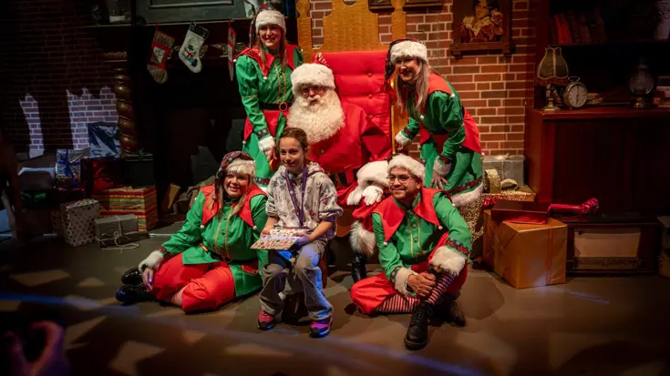 De kerstman poseert met een kindje voor een fotograaf, omgeven door helpers van de kerstman.