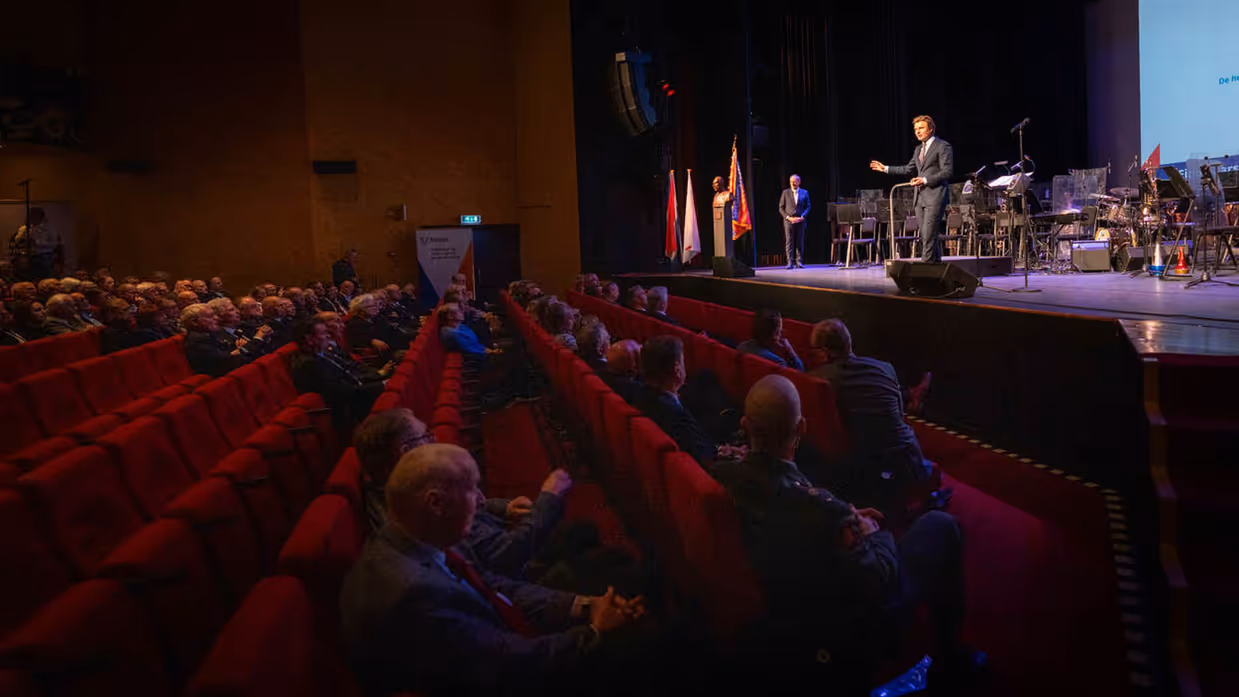 Minister Ruben Brekelmans op het podium, voor hem rode stoelen in het publiek met mensen erop.