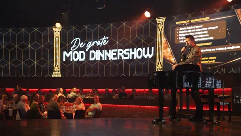 Een zaal vol mensen kijkt naar een man op een podium. Op de achtergrond is een billboard te zien met de tekst ‘De Grote MOD-dinnershow’.