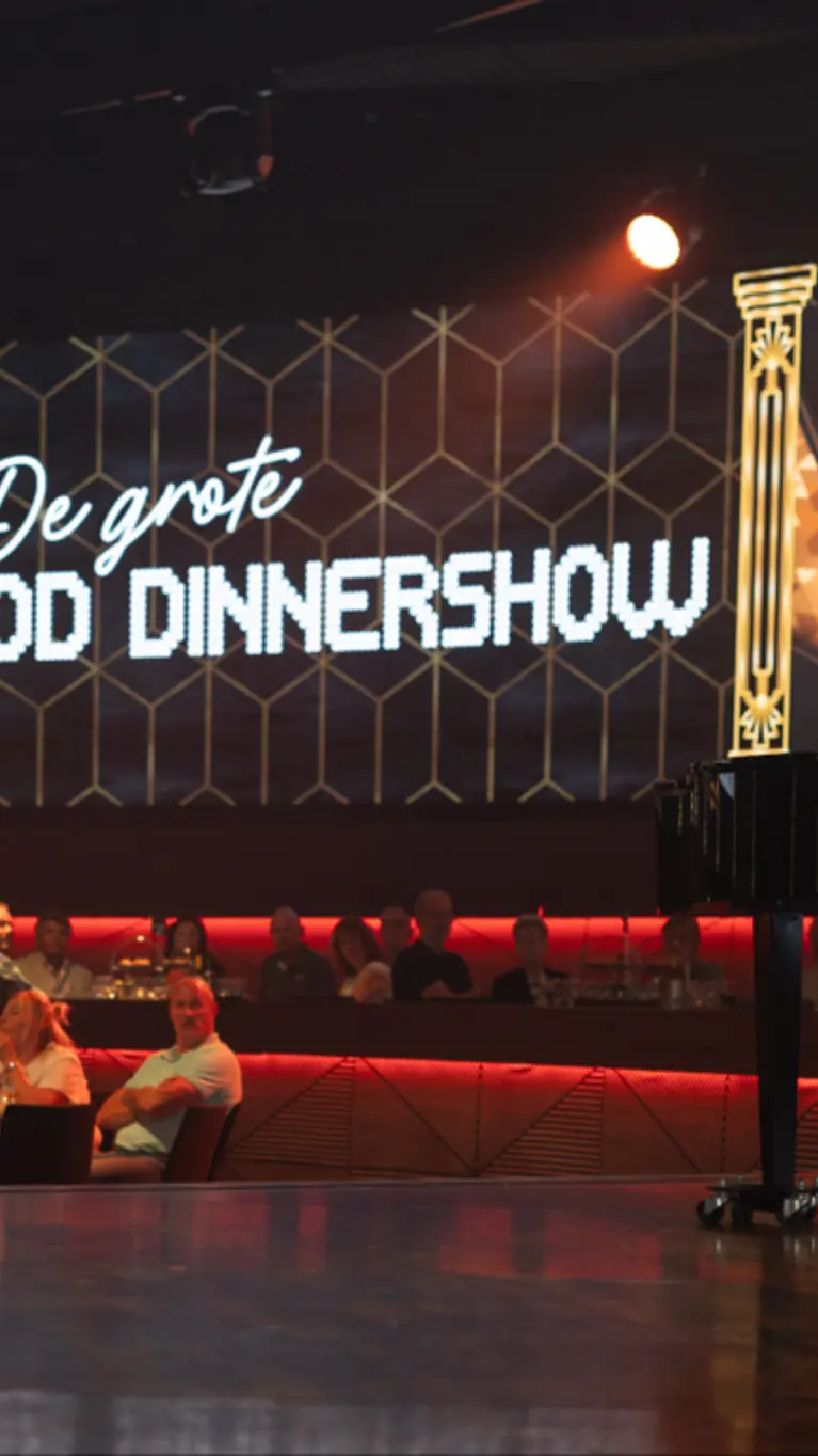 Een zaal vol mensen kijkt naar een man op een podium. Op de achtergrond is een billboard te zien met de tekst ‘De Grote MOD-dinnershow’.