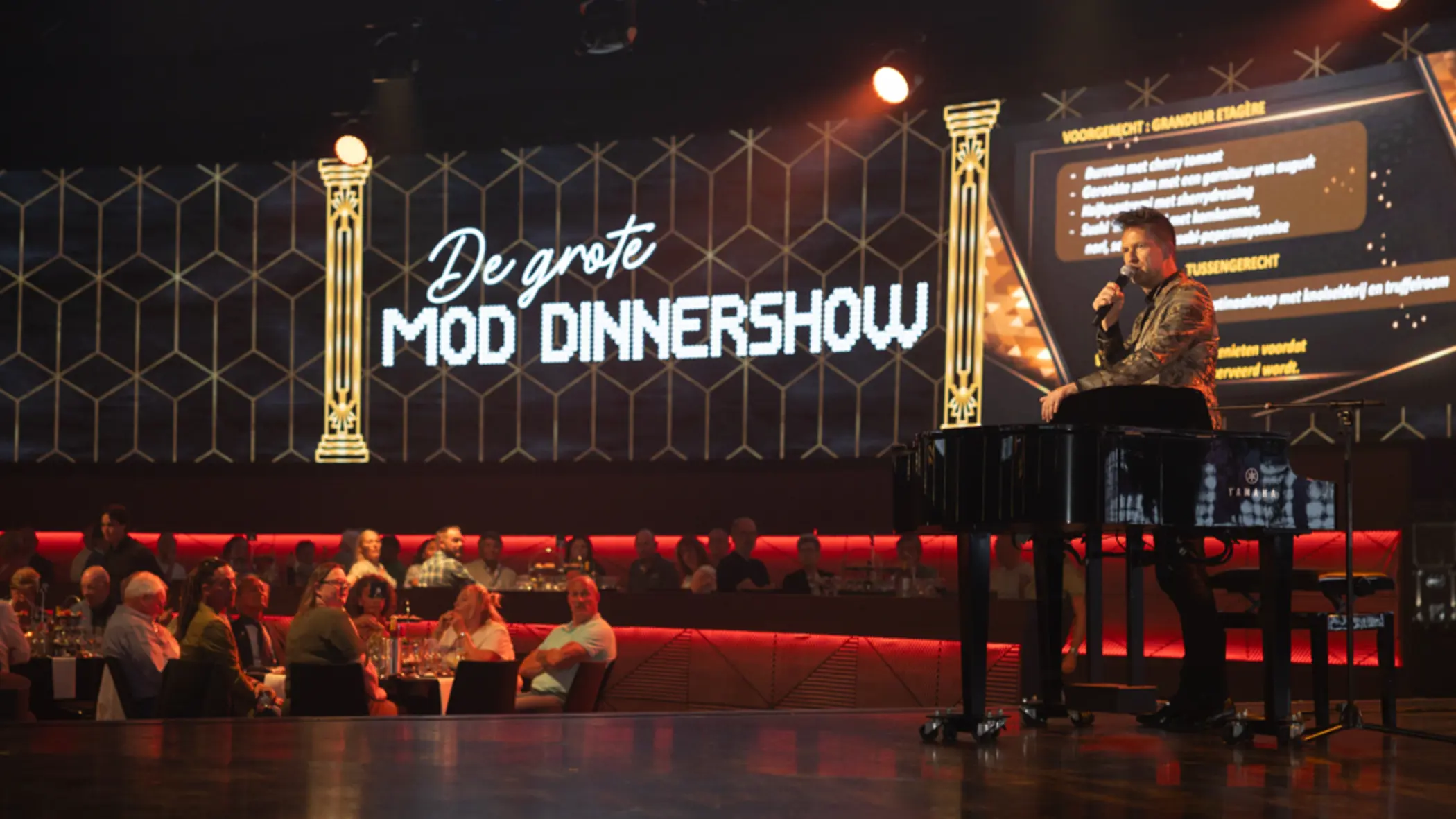 Een zaal vol mensen kijkt naar een man op een podium. Op de achtergrond is een billboard te zien met de tekst ‘De Grote MOD-dinnershow’.