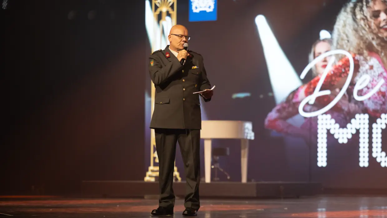 John staat in DT-tenue op het podium met een microfoon in zijn hand. 