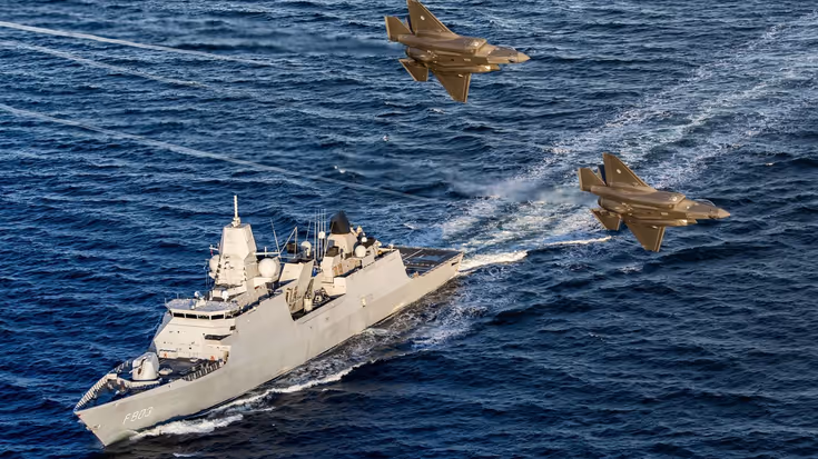 Een marineschip op zee met daarboven 2 gevechtsvliegtuigen.