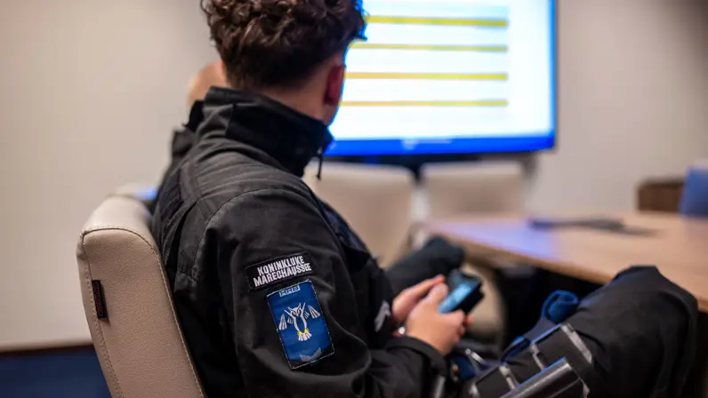 Een cursist van de Koninklijke Marechaussee volgt een briefing terwijl routes en aandachtspunten op een scherm worden besproken.