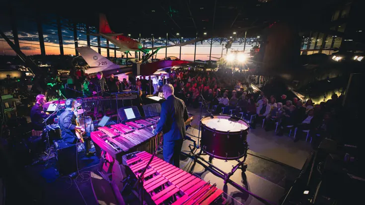 Militair orkest in sfeervol verlichte ruimte speelt muziek voor een publiek dat tussen vliegtuigen zit.