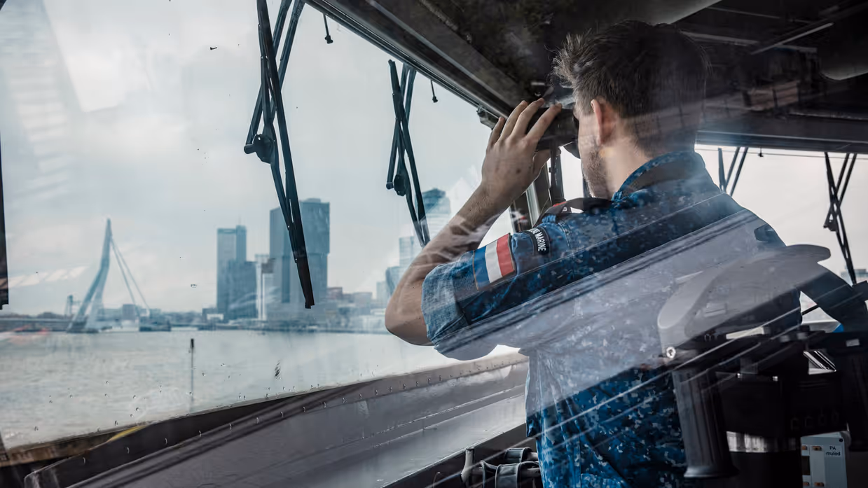 Marineman kijkt vanaf brug van het schip door verrekijker richting Erasmusbrug.