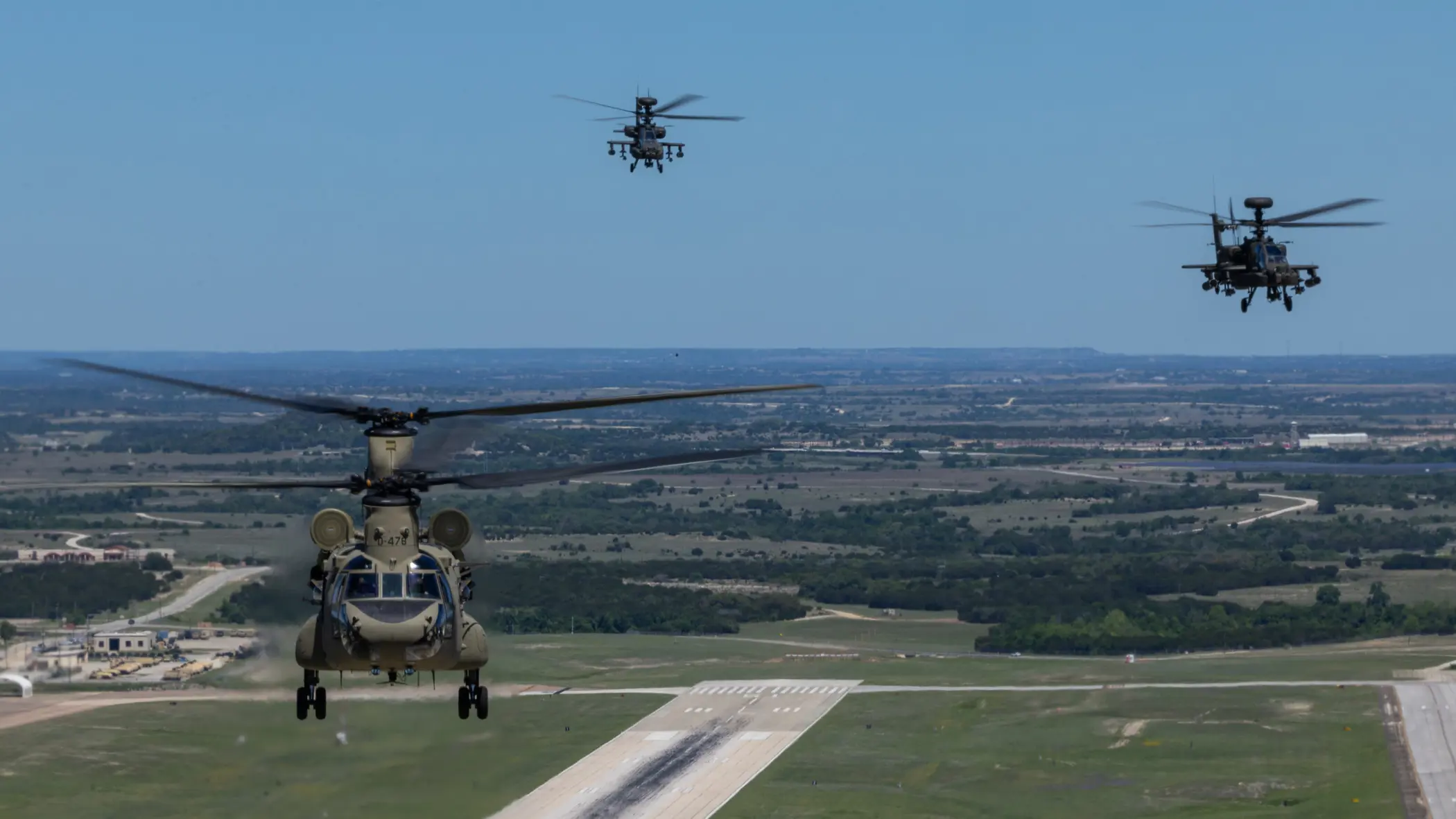 Een Chinook en twee Apaches vliegend boven een startbaan met op de achtergrond een blauwe lucht en weids uitzicht over vlak platteland.