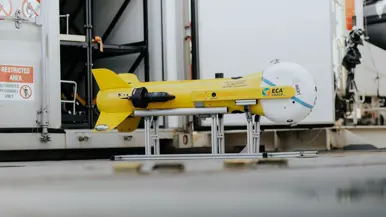 Links een grijs bootje met achterop verschillende drones. Dit vaart langs een groot vrachtschip. Daarnaast een gele, raketvormige drone met voorop een witte bol. Deze is ook te zien achterop het varende bootje.