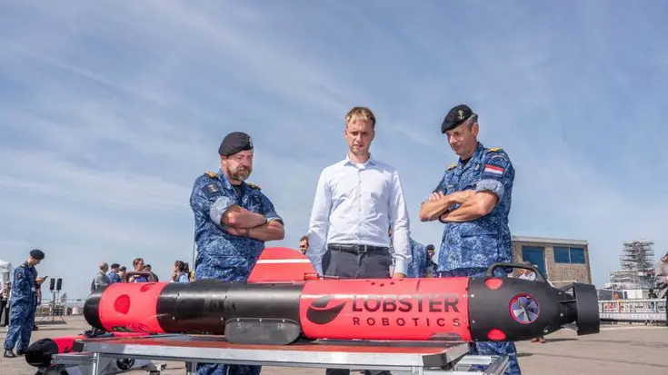 Twee militairen en een man in burgerkleding bekijken drone die voor hen op tafel ligt.