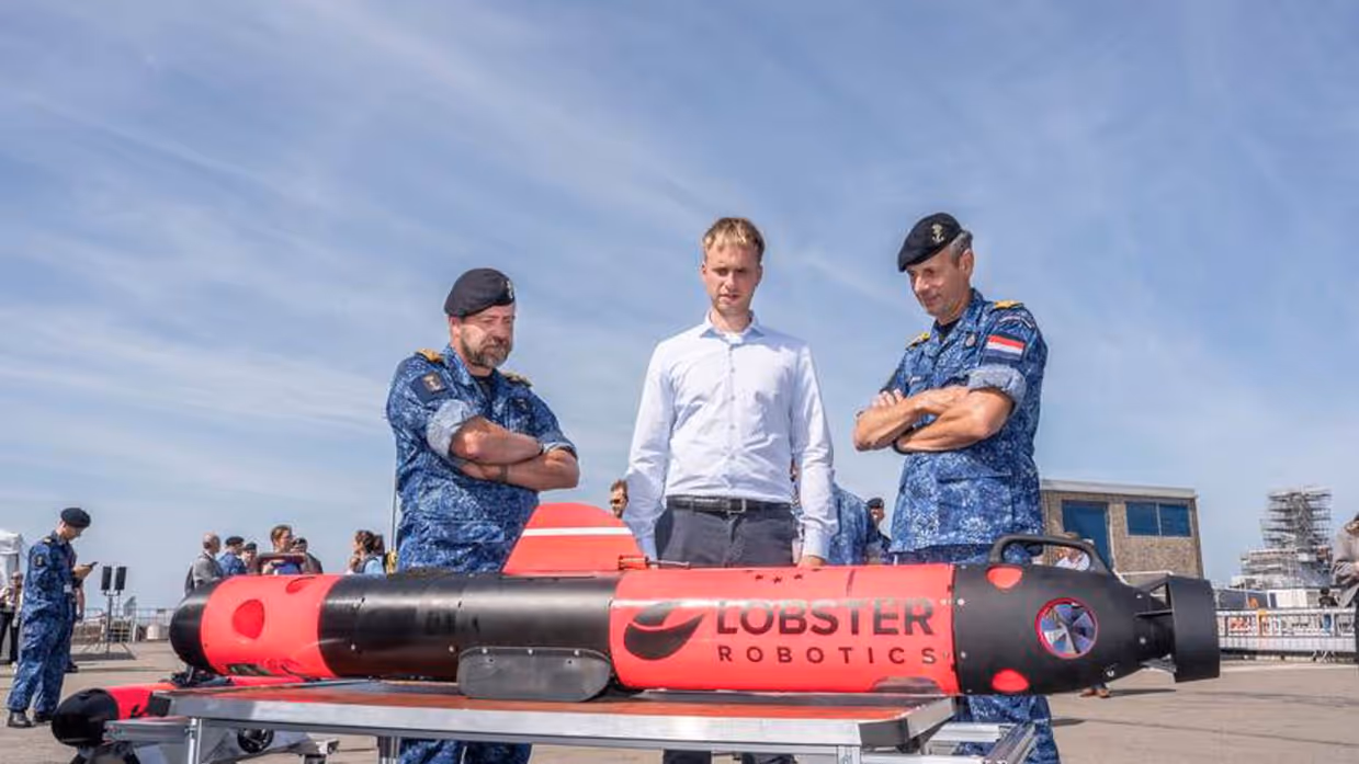 Twee militairen en een man in burgerkleding bekijken drone die voor hen op tafel ligt.