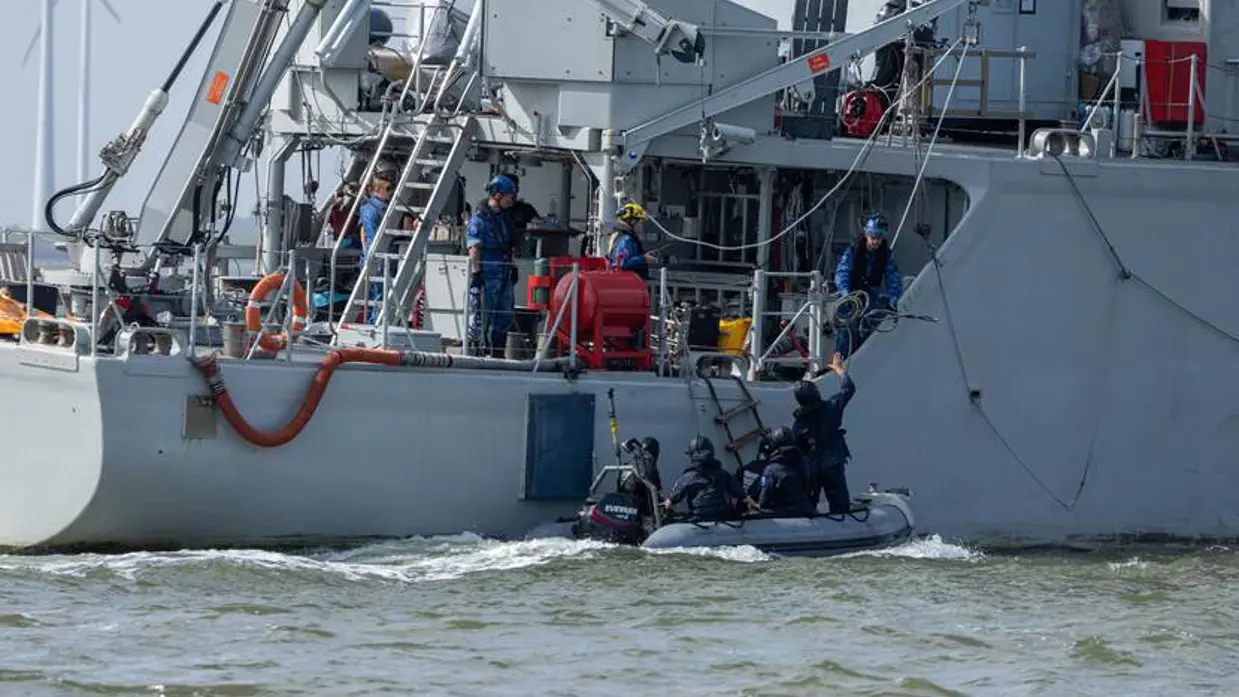 Vier zwartgeklede mensen in een rubberboot liggen naast een grijs schip bij een touwladder.