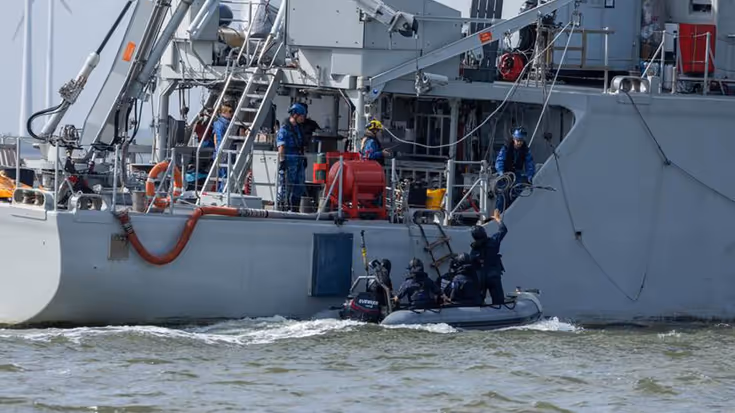 Vier zwartgeklede mensen in een rubberboot liggen naast een grijs schip bij een touwladder.