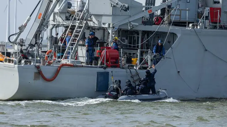 Vier zwartgeklede mensen in een rubberboot liggen naast een grijs schip bij een touwladder.