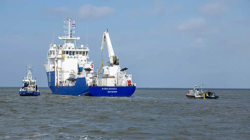 Het schip de NS Geosea, van de onderkant blauw en de bovenkant wit, vaart op zee. Links en rechts een klein bootje.