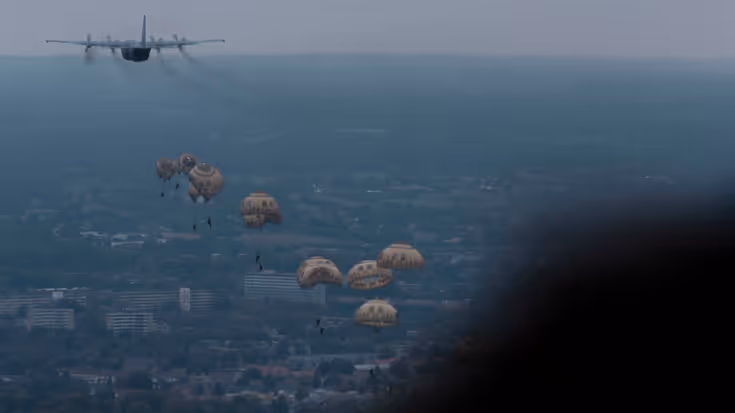 Parachutisten hangen aan hun parachutes onderweg naar beneden. Linksboven het vliegtuig waar ze net uit zijn gesprongen. 