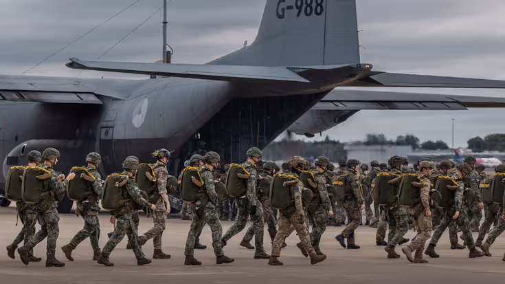 Hercules met op de voorgrond militairen die met een parachute op hun rug lopen. 