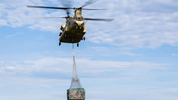 Een Chinook met in de lucht met Bambi Bucket die eronder hangt.