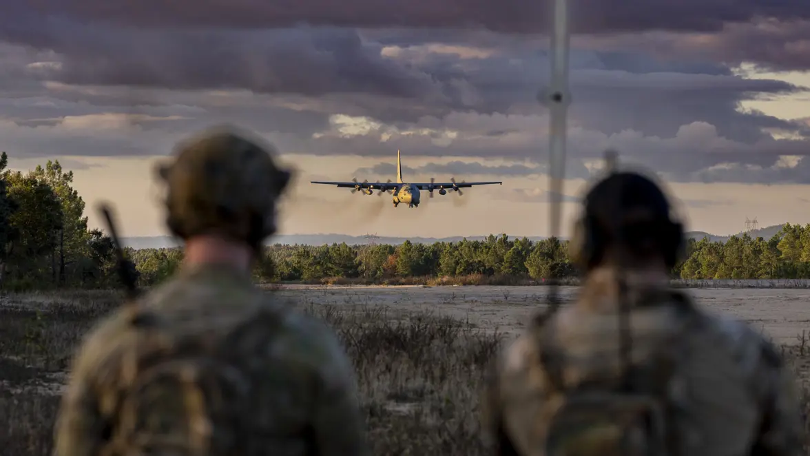 Een C-130 Hercules nadert een open vlakte, op de voorgrond twee militairen die vanuit de bosrand toekijken.