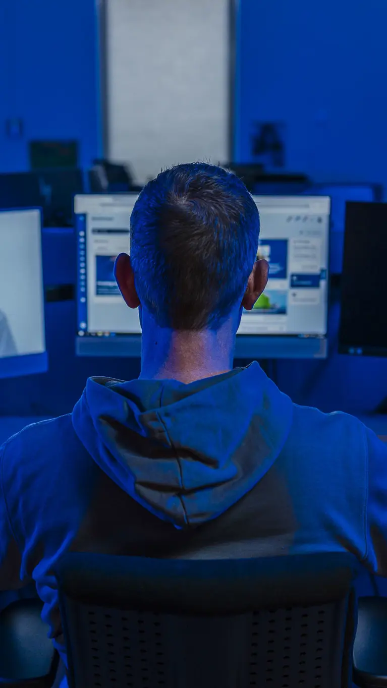 Professionele werkomgeving met een persoon aan een bureau, kijkend naar meerdere computerschermen in een blauw verlichte ruimte. Ook een silhouet van een gemaskerde figuur op één van de schermen.