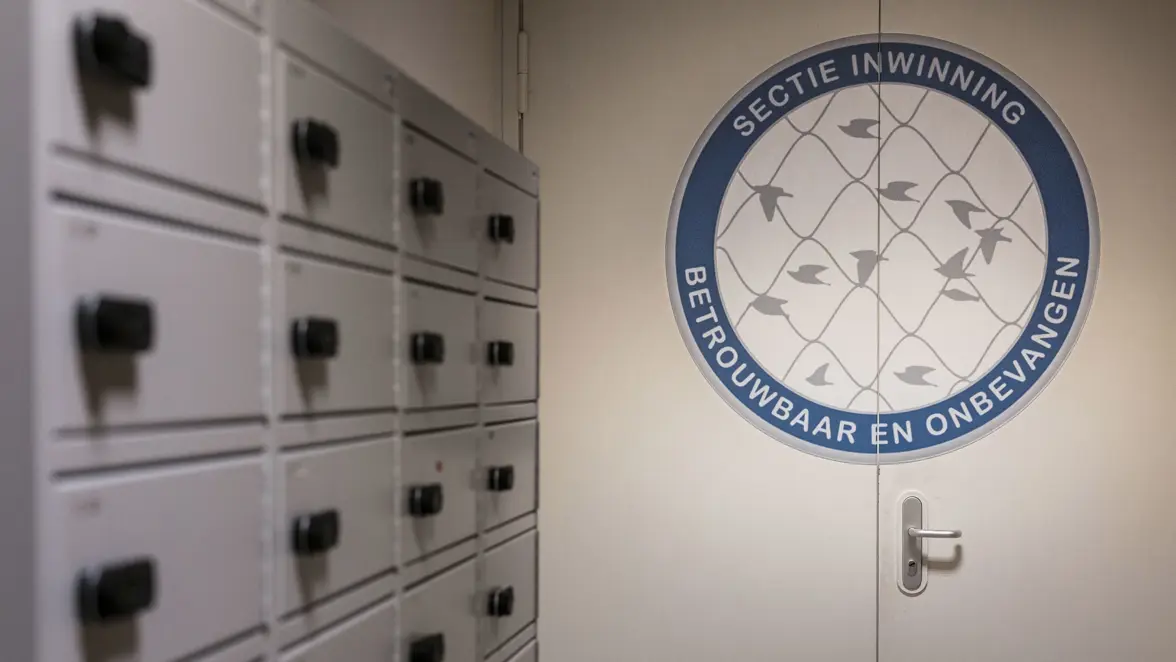 Gang met lockers naast een deur met een groot rond embleem waarop staat “Sectie Inwinning. Betrouwbaar en Onbevangen” en een patroon van vliegende vogels.