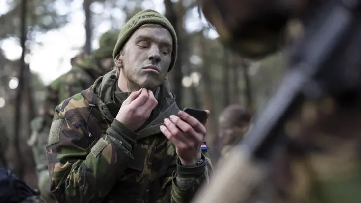 Een deelnemer brengt camouflagekleuren op op zijn gezicht. Hij heeft 1 hand bij zijn kin.