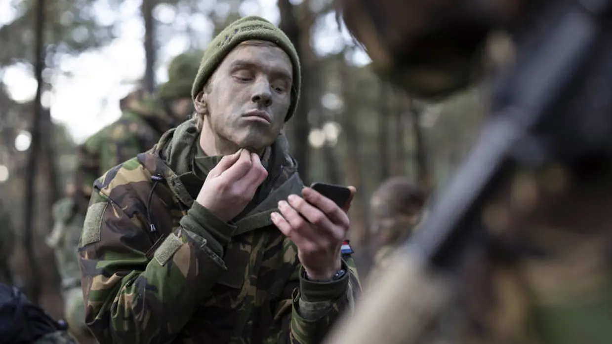 Een deelnemer brengt camouflagekleuren op op zijn gezicht. Hij heeft 1 hand bij zijn kin.
