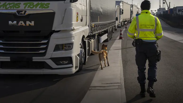 Marechaussee met hond op de rug gezien loopt langs trailer.