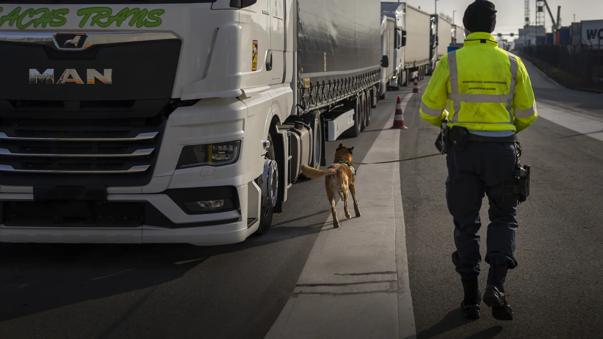 Marechaussee met hond op de rug gezien loopt langs trailer.