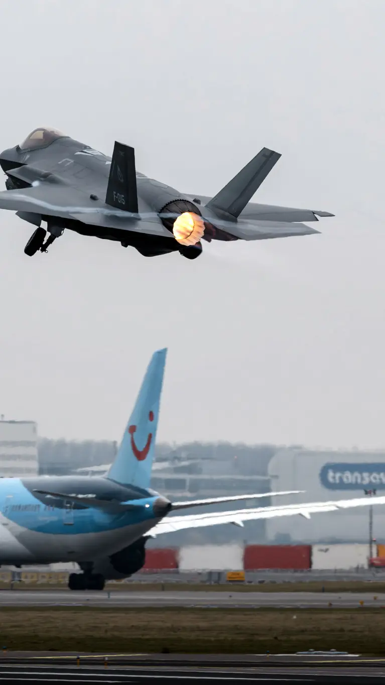 Een F-35 gevechtsvliegtuig stijgt op vanaf luchthaven Schiphol. Op de achtergrond is de luchthaven en een taxiënd civiel passagiersvliegtuig te zien.