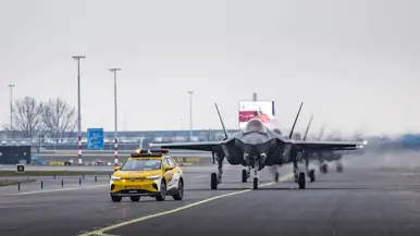 Een geparkeerde F-35 staat op luchthaven Schiphol, met de klep van de cockpit geopend. Om het toestel bewegen militairen, die onderhoud plegen. Op de voorgrond loopt iemand met een schijf met de tekst ‘remove before flight’. De luchtmachters dragen een groene militaire broek, met daarboven een oranje veiligheidshesje. Daarnaast staat een foto van taxiende F-35’s op het vliegveld. Op de achtergrond is de snelweg te zien.