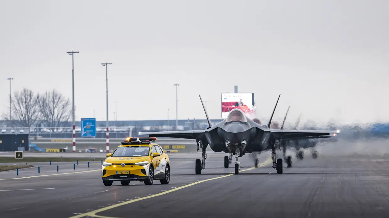 Een geparkeerde F-35 staat op luchthaven Schiphol, met de klep van de cockpit geopend. Om het toestel bewegen militairen, die onderhoud plegen. Op de voorgrond loopt iemand met een schijf met de tekst ‘remove before flight’. De luchtmachters dragen een groene militaire broek, met daarboven een oranje veiligheidshesje. Daarnaast staat een foto van taxiende F-35’s op het vliegveld. Op de achtergrond is de snelweg te zien.