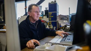 Een medewerker monitort van dichtbij hoe een pallet wordt uitgeladen. Op de tweede foto een medewerker, gezeten achter zijn bureau.