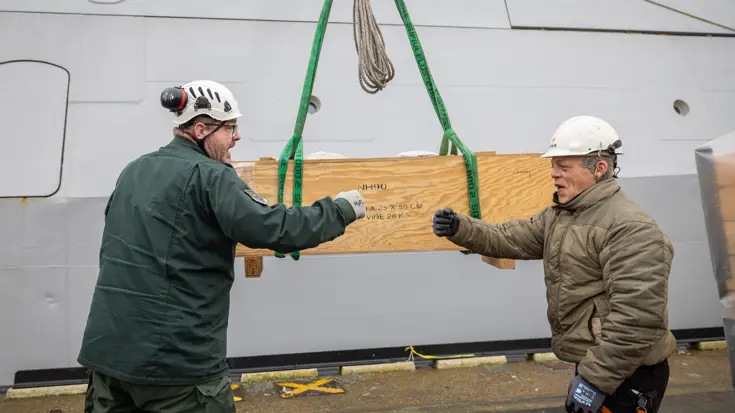 Twee medewerkers geven elkaar een boks tegen de achtergrond van een houten kist met daarop de tekst NH90.