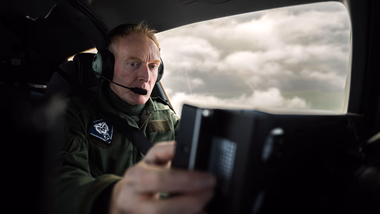 Een piloot zit in de cockpit van het patrouillevliegtuig achter het stuur, rechts van hem is een raam met uitzicht op het wolkendek.
