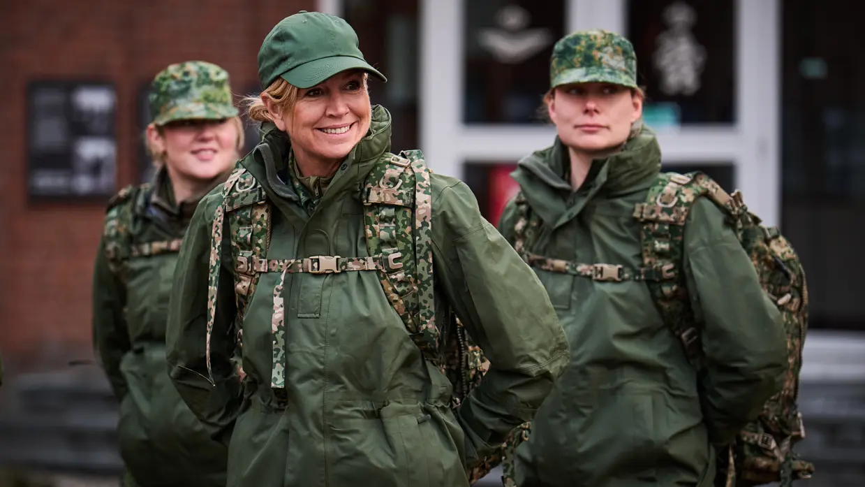 Een lachende koningin Máxima in gevechtstenue met pet op en rugzak om, achter haar twee vrouwelijke militairen in dezelfde outfit.