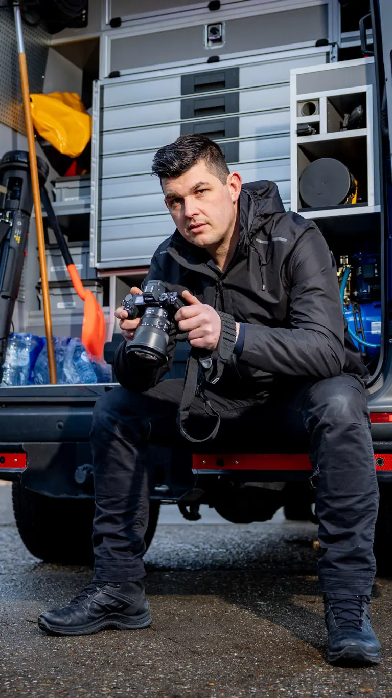 Adjudant Allard zit achterin de dienstbus met zijn camera vast en een blik naar de camera toe.