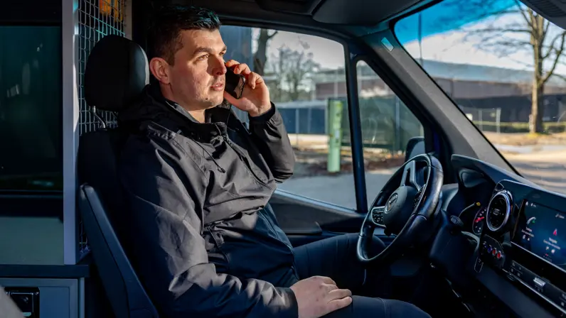 Adjudant Allard op de bestuurdersstoel van de dienstbus, met een mobiele telefoon aan zijn oor.