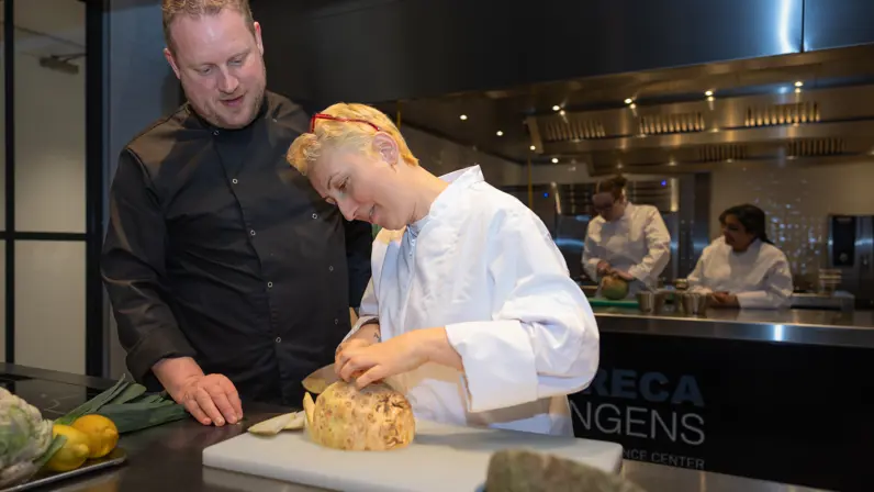 Dijan staat in een keuken en snijdt onder toeziend oog van een docent een knol. 