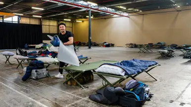 Van links naar rechts: een jongeman pakt iets van een veldbedje met een slaapzaak in een grote zaal. Een militair in een blauw uniform legt iets uit aan een groepje toehoorders in een donkere zaal. Twee man zitten aan een schaakbord met een poster van Defensie op de achtergrond. Een man in uniform met strepen op de schouder in bespreking met vijf anderen.