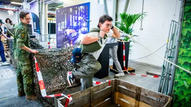 Diverse beelden van de fysieke uitdagingen, van links naar rechts: een team loopt met de handen aan een touw, waarbij ze over hindernissen moeten komen. Een team is gemaskerd en moet een ei in een dop over een parcours leiden. Rechts hangen twee deelnemers aan een rekstok.