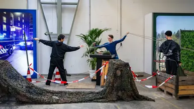 Diverse beelden van de fysieke uitdagingen, van links naar rechts: een team loopt met de handen aan een touw, waarbij ze over hindernissen moeten komen. Een team is gemaskerd en moet een ei in een dop over een parcours leiden. Rechts hangen twee deelnemers aan een rekstok.