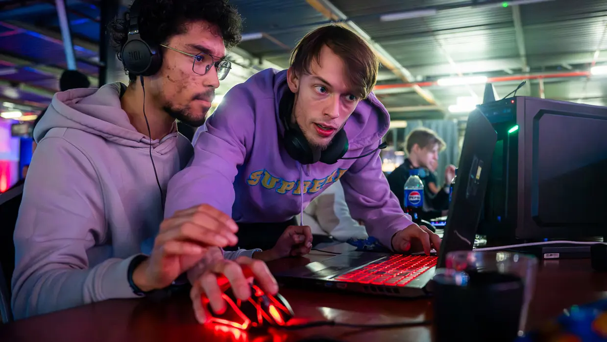 Van links naar rechts: in een zaal met veel tafels sluit een jongen zijn apparatuur aan op een computer, die lichtgevend op het bureau staat. Een man lijkt aanwijzingen te geven aan een gamer achter de computer, dan geven ze elkaar een high five.