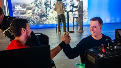 Van links naar rechts: in een zaal met veel tafels sluit een jongen zijn apparatuur aan op een computer, die lichtgevend op het bureau staat. Een man lijkt aanwijzingen te geven aan een gamer achter de computer, dan geven ze elkaar een high five.