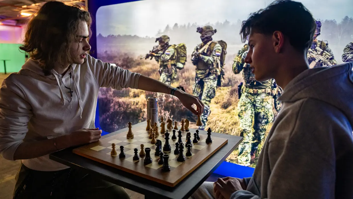 Van links naar rechts: een jongeman pakt iets van een veldbedje met een slaapzaak in een grote zaal. Een militair in een blauw uniform legt iets uit aan een groepje toehoorders in een donkere zaal. Twee man zitten aan een schaakbord met een poster van Defensie op de achtergrond. Een man in uniform met strepen op de schouder in bespreking met vijf anderen.