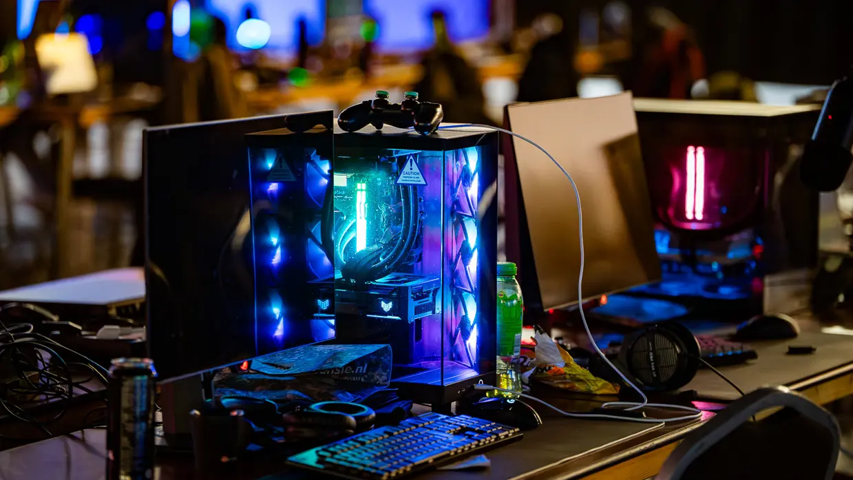 Van links naar rechts: in een zaal met veel tafels sluit een jongen zijn apparatuur aan op een computer, die lichtgevend op het bureau staat. Een man lijkt aanwijzingen te geven aan een gamer achter de computer, dan geven ze elkaar een high five.
