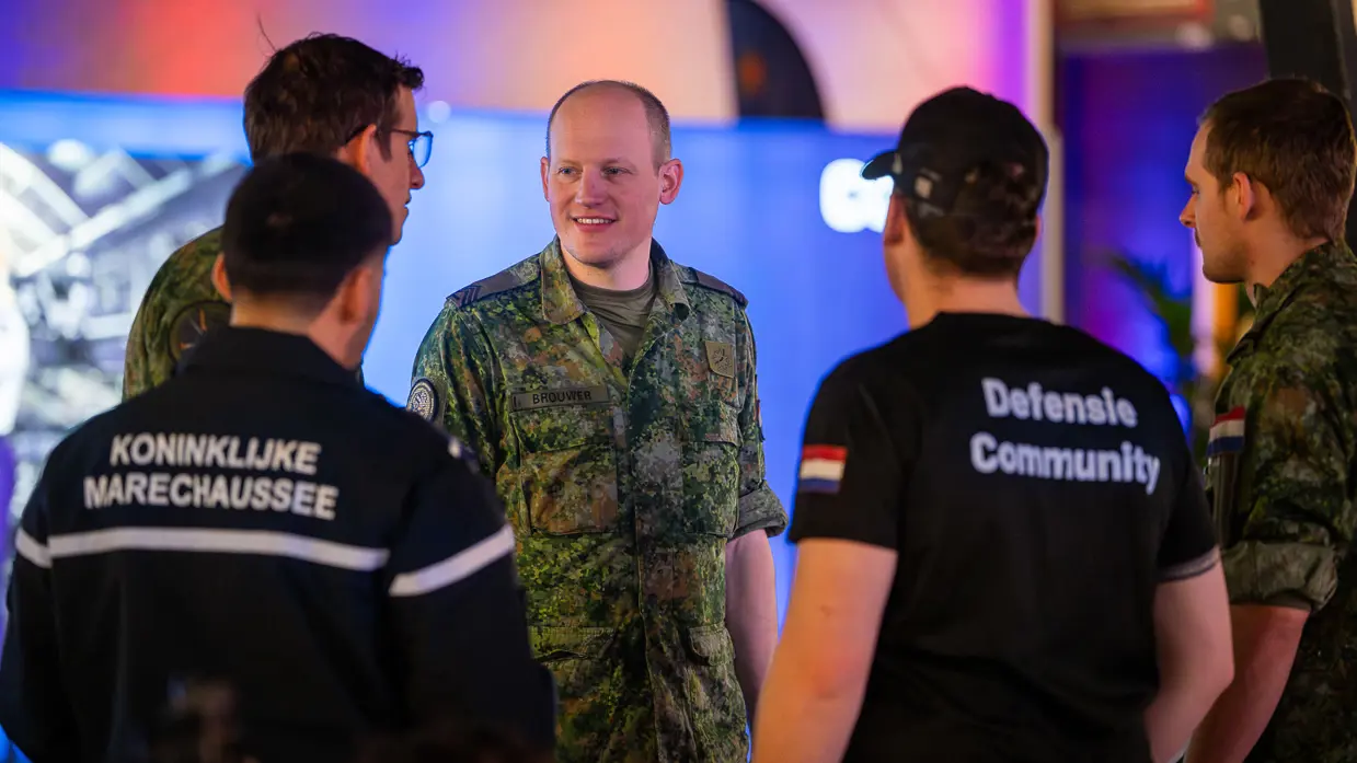 Van links naar rechts: een jongeman pakt iets van een veldbedje met een slaapzaak in een grote zaal. Een militair in een blauw uniform legt iets uit aan een groepje toehoorders in een donkere zaal. Twee man zitten aan een schaakbord met een poster van Defensie op de achtergrond. Een man in uniform met strepen op de schouder in bespreking met vijf anderen.
