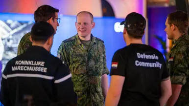 Van links naar rechts: een jongeman pakt iets van een veldbedje met een slaapzaak in een grote zaal. Een militair in een blauw uniform legt iets uit aan een groepje toehoorders in een donkere zaal. Twee man zitten aan een schaakbord met een poster van Defensie op de achtergrond. Een man in uniform met strepen op de schouder in bespreking met vijf anderen.