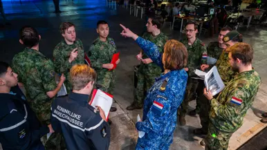 Van links naar rechts: een jongeman pakt iets van een veldbedje met een slaapzaak in een grote zaal. Een militair in een blauw uniform legt iets uit aan een groepje toehoorders in een donkere zaal. Twee man zitten aan een schaakbord met een poster van Defensie op de achtergrond. Een man in uniform met strepen op de schouder in bespreking met vijf anderen.
