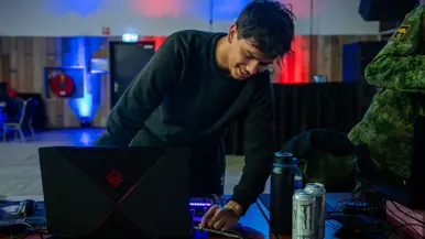 Van links naar rechts: in een zaal met veel tafels sluit een jongen zijn apparatuur aan op een computer, die lichtgevend op het bureau staat. Een man lijkt aanwijzingen te geven aan een gamer achter de computer, dan geven ze elkaar een high five.