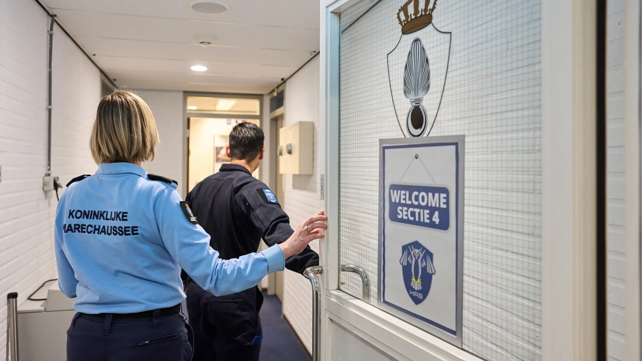 Adjudant Carolien en kapitein Tobias stappen door een deur waarop ‘Welcome Sectie 4’ wordt afgebeeld.